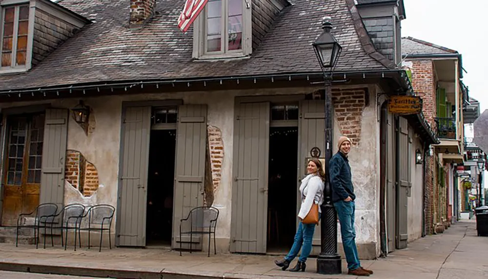 Two people are standing in front of a quaint building with a curved shingled roof on a historic street possibly in the French Quarter of New Orleans