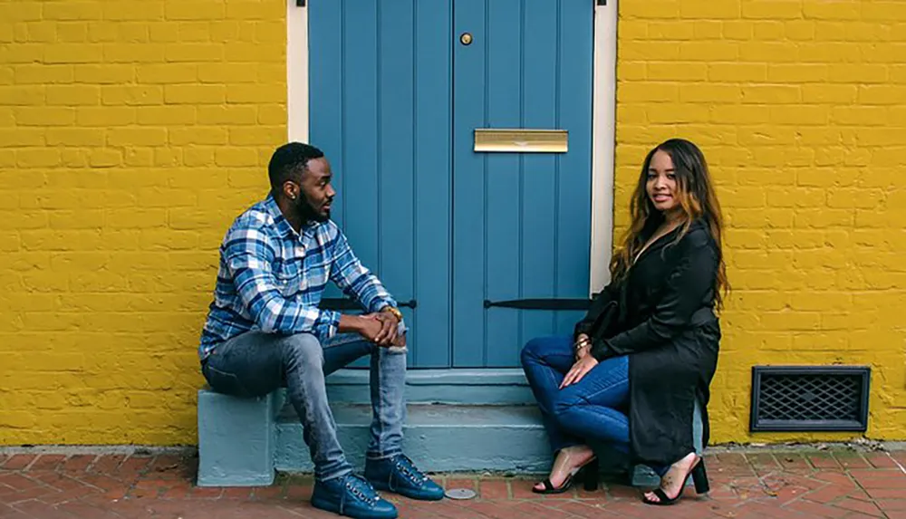 A man and a woman are sitting apart on a doorstep and bench against a yellow wall with a blue door appearing to be in a moment of casual contemplation or conversation