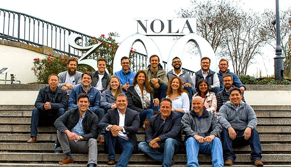 A group of smiling people are posing for a photo on steps in front of a large sign that reads NOLA ZOO