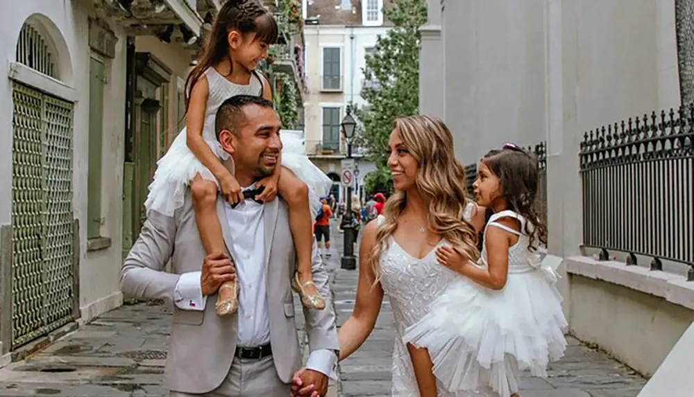 A family is walking down a city street with a smiling man carrying a young girl on his shoulders holding hands with a woman in a white dress while another girl walks beside them