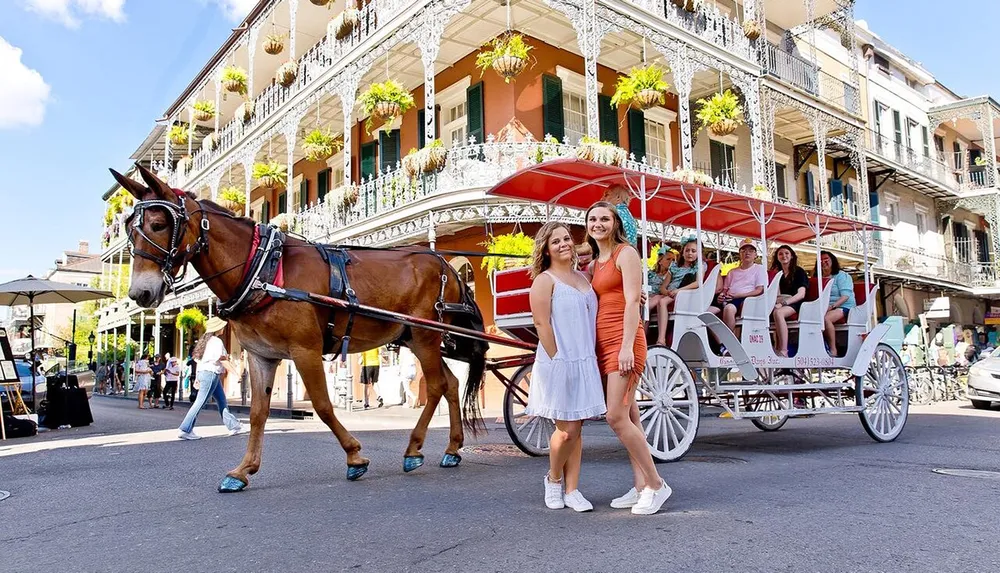 A horse-drawn carriage waits on a lively street with passengers as two women pose for a photo in the foreground set against the backdrop of a balcony-adorned building