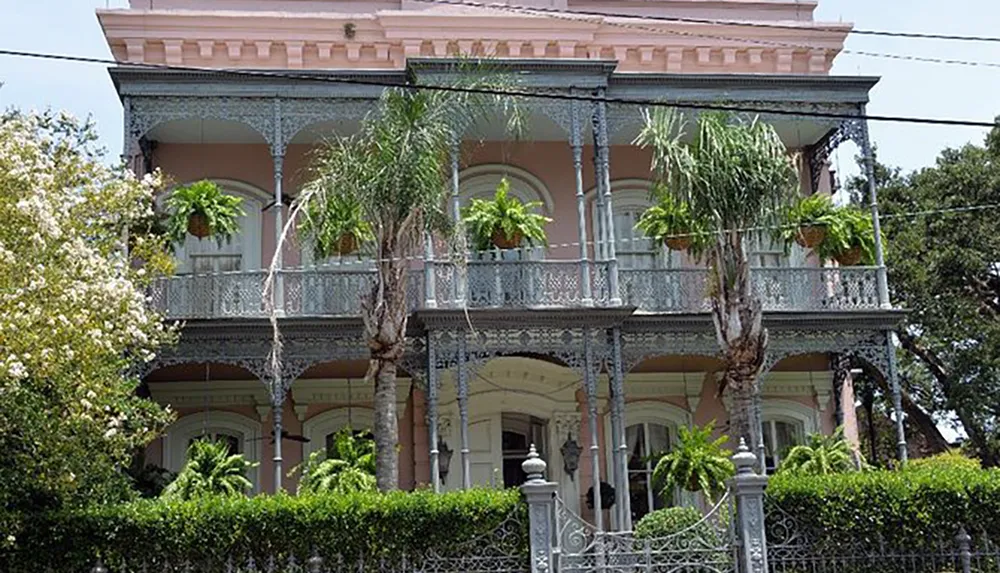 The image shows a two-story pastel-colored house with intricate ironwork on the balconies surrounded by lush greenery and a decorative iron fence