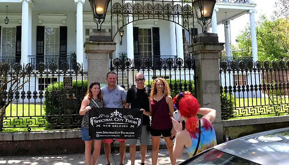 A group of tourists is posing for a photo with a Spectral City Tours sign in front of an ornate gated building likely suggesting they are on a ghost or historical tour in New Orleans