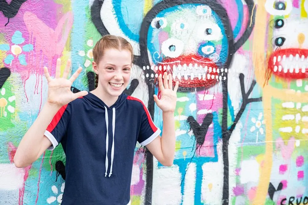 A smiling girl is posing with her hands up in front of a vibrant colorful graffiti wall