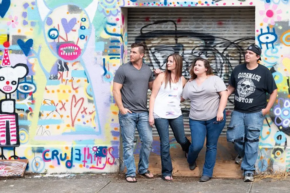 Four individuals are standing in front of a colorful graffiti-covered wall posing casually and looking away from the camera