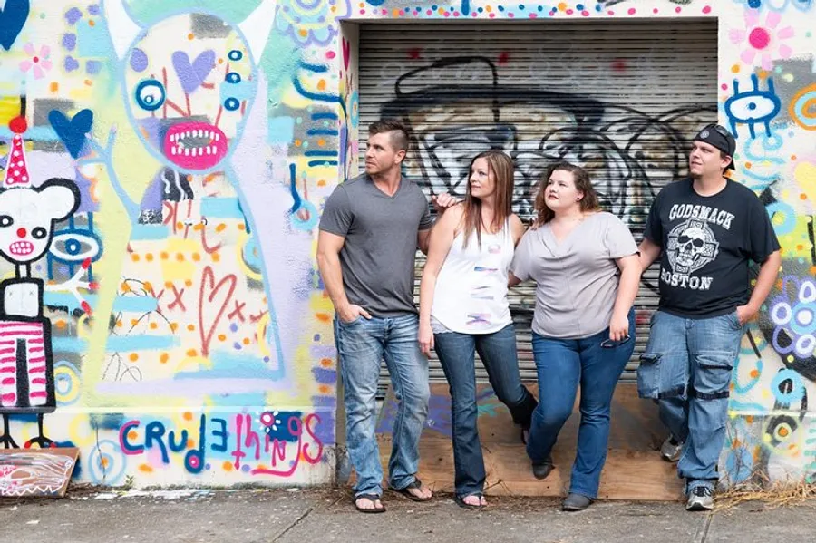 Four individuals are standing in front of a colorful, graffiti-covered wall, posing casually and looking away from the camera.