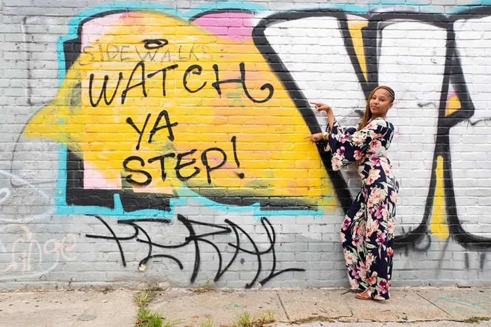 A person is standing against a graffiti-covered wall with a phrase WATCH YA STEP painted on it and they are pointing towards the text