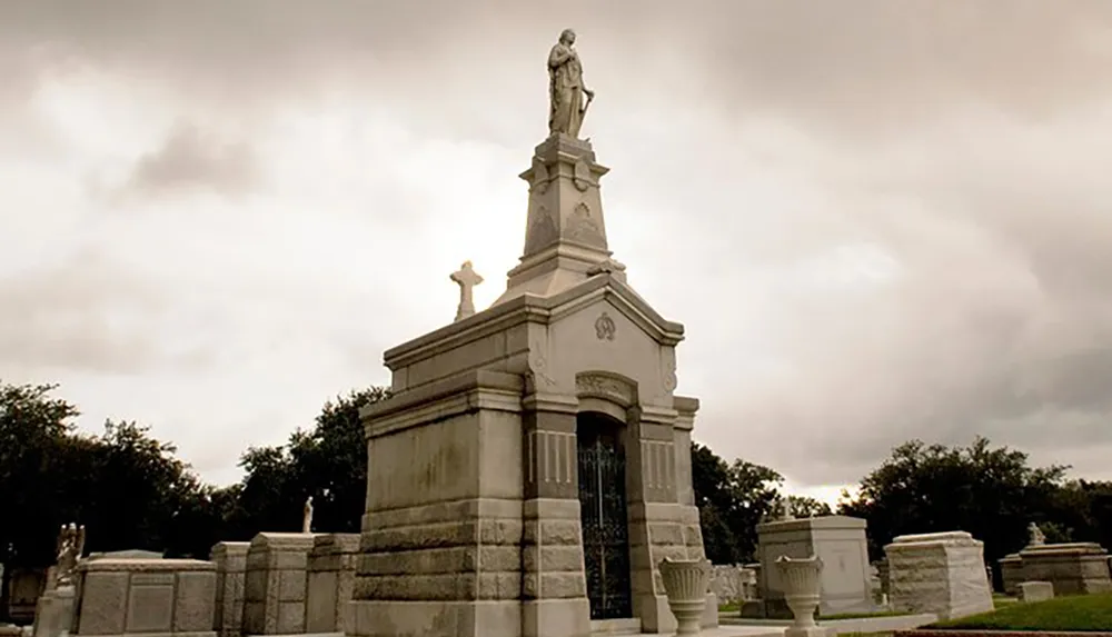 The image features an ornate mausoleum with a statue atop situated under a cloudy sky in a cemetery dotted with similar structures