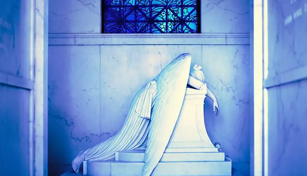 The image shows a sorrowful angelic statue draped over a pedestal in a room with blue hues and a stained glass window