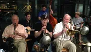 A group of musicians performs live, with instruments like a clarinet, trumpet, trombone, guitar, and a double bass, creating a scene of classic, possibly jazz, ensemble.