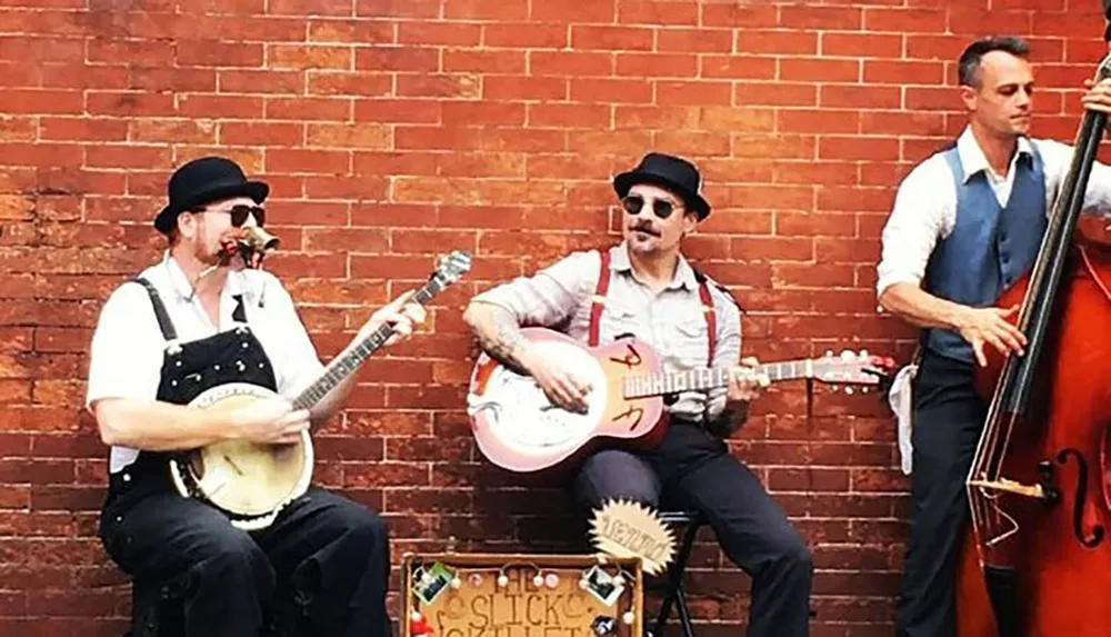 Two musicians are playing a banjo and a guitar while sitting and a third is standing playing an upright bass against a brick wall background likely performing live music on the street