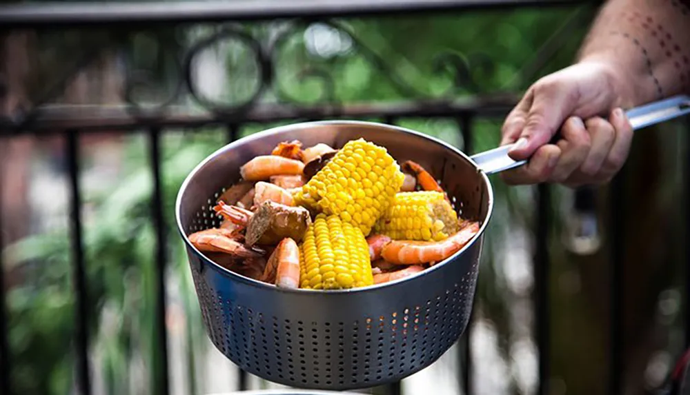 A person is holding a metal colander filled with boiled seafood including shrimp and corn on the cob potentially indicating a seafood boil meal