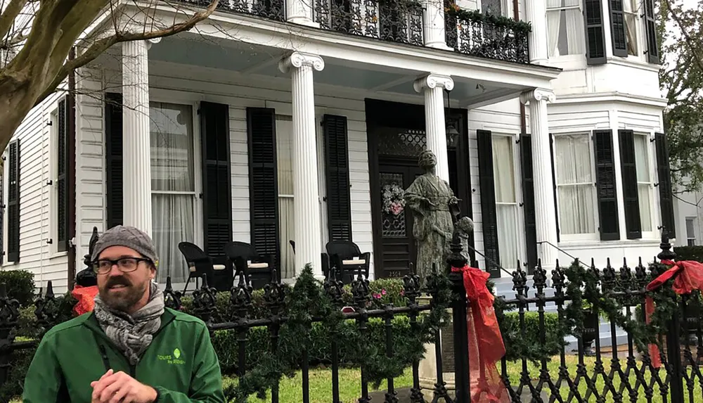 A man in a green jacket stands in front of a classic white house with black shutters ornate ironwork and a statue in the yard