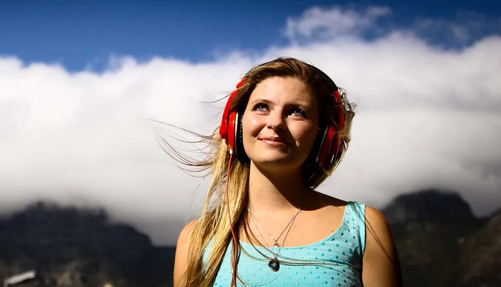 A young woman is wearing red headphones and smiling while looking up towards the sky with clouds and mountains in the background