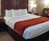 The image shows a well-made bed with white linens and a red decorative runner in a tidy neutrally decorated hotel room
