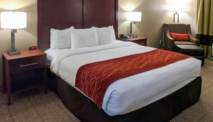 The image shows a well-made bed with white linens and a red decorative runner in a tidy neutrally decorated hotel room