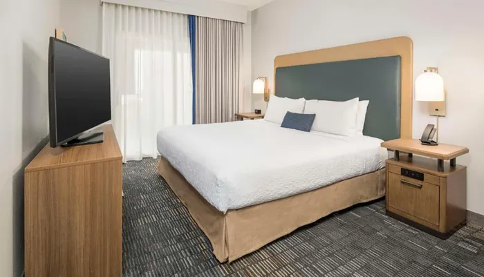 The image shows a tidy and modern hotel room with a large bed a flat-screen TV and a window with curtains