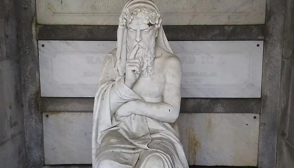 The image shows a marble sculpture of a bearded seated male figure likely representing a philosophical or mythological character in a contemplative pose with a finger to his lips