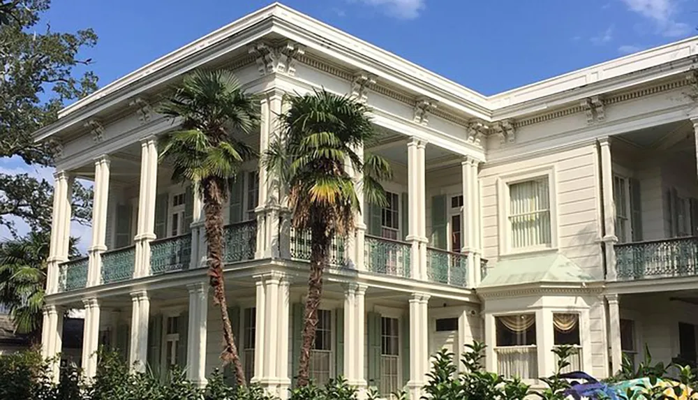 The image shows a grand two-story house with a classical architectural style featuring tall columns balconies with ornate railings and lush surrounding vegetation under a blue sky