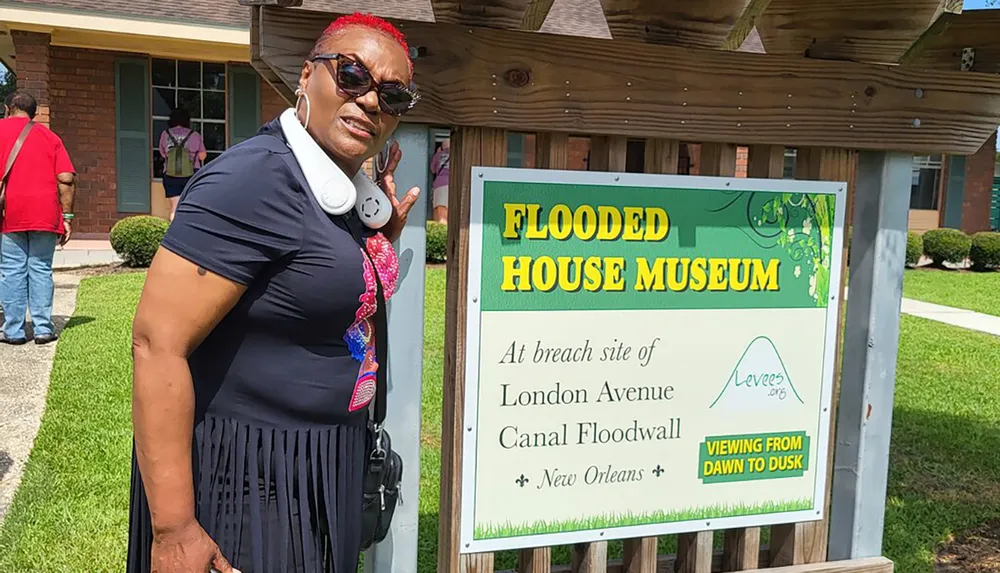 A woman is posing next to a sign that reads Flooded House Museum at the breach site of London Avenue Canal Floodwall in New Orleans