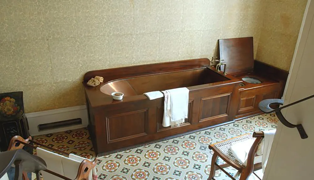 The image shows an antique wooden bathtub with integrated basin and toilet in a vintage-styled bathroom with patterned tile flooring
