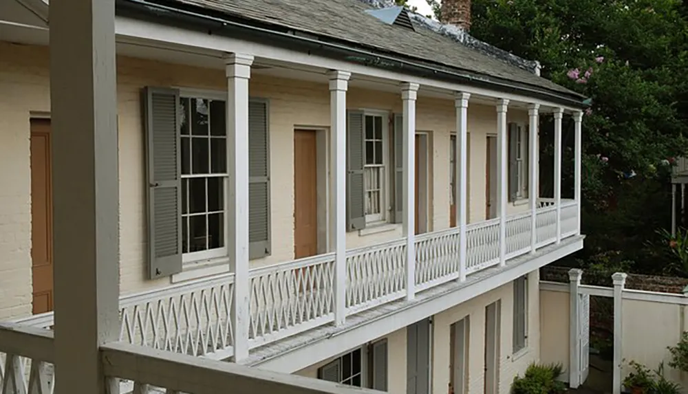 The image shows the side of an old building with a covered wooden balcony featuring white railings and columns set against a backdrop of green foliage