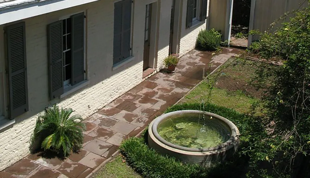 This image shows a courtyard with a small circular fountain in the center bordered by wet flagstone paths green shrubs and a building with closed shutters
