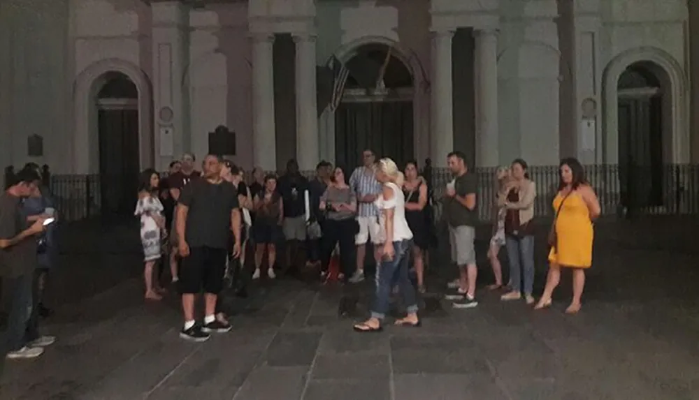 A group of people are standing outside at night possibly on a tour with some individuals looking towards a central point of interest as others engage with their mobile devices