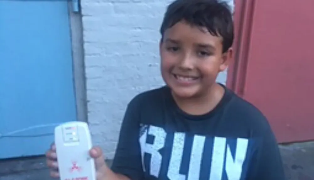 A young boy is smiling at the camera holding a white electronic device with a red cross on it standing in front of a blue door and a brick wall