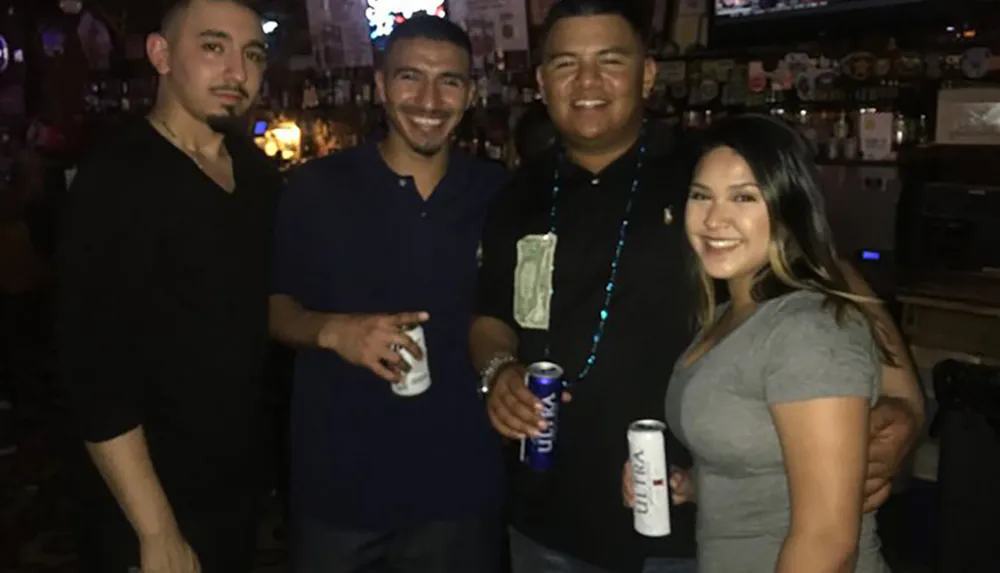 Four people are smiling for a photo while standing together in a bar setting with some holding cans of beer