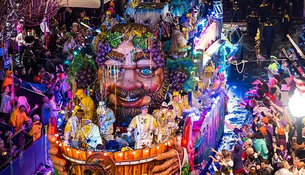 A vibrant and crowded street parade features individuals in colorful costumes on a float with a large whimsical character head surrounded by revelers