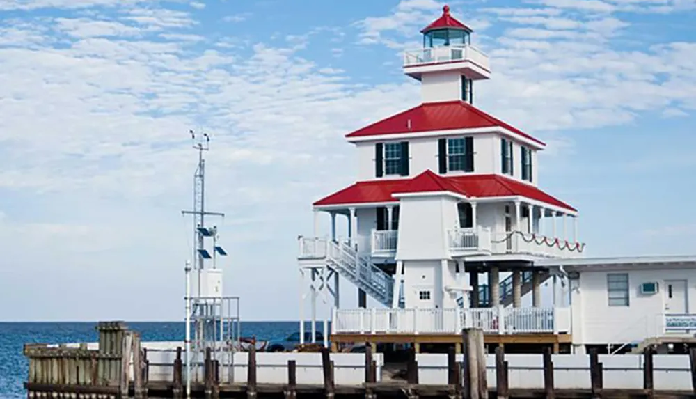 A picturesque red-and-white lighthouse stands on a wooden pier overlooking the sea under a sky dotted with clouds