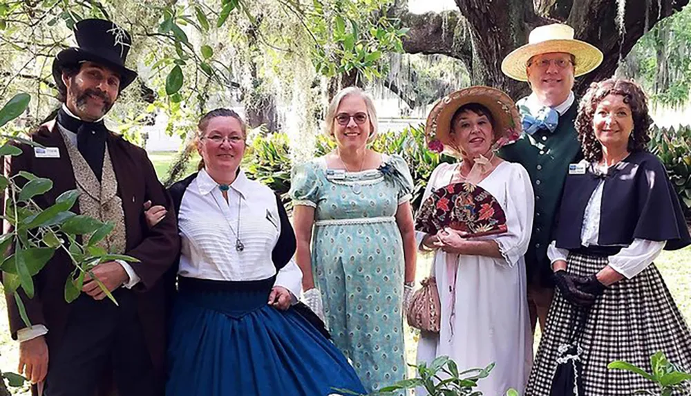 Six individuals are dressed in historical clothing reminiscent of the 19th century posing outdoors for a themed event or reenactment