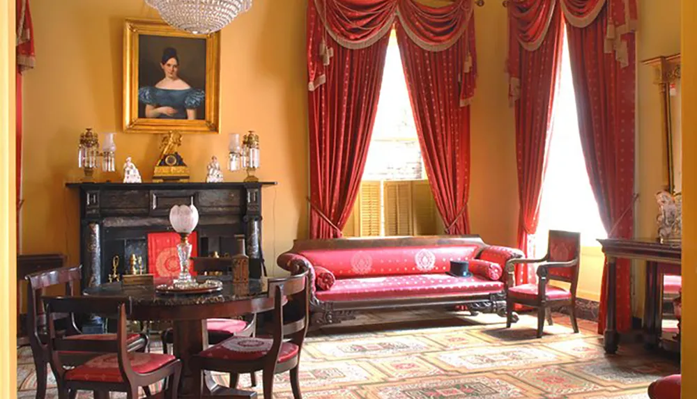 The image showcases a stately room with a classic Victorian-era decor featuring a red upholstered sofa matching drapes antique wooden furniture a chandelier and a framed portrait above the fireplace