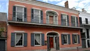 The image shows a two-story red brick building with a balcony, gray shutters, and a white cornice, reflecting a typical architectural style found in the French Quarter of New Orleans.