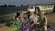 A group of six people posing with bicycles adorned with colorful LED lights by a riverfront at dusk.