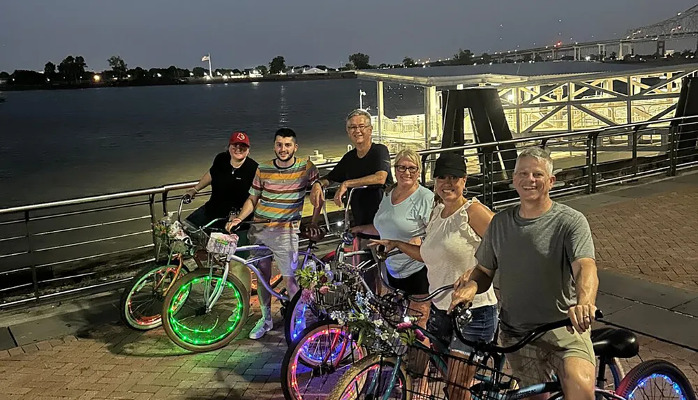 A group of six people posing with bicycles adorned with colorful LED lights by a riverfront at dusk
