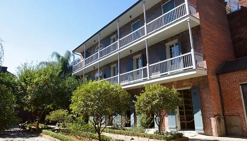 The image shows a two-story brick building with white balconies surrounded by green shrubbery under a clear blue sky