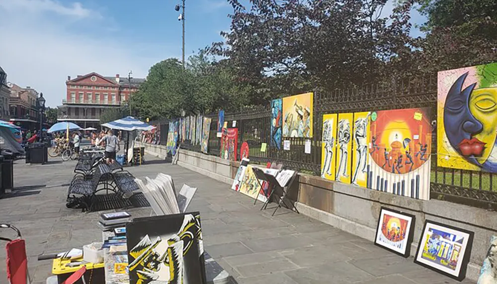 An outdoor art market displays a variety of colorful paintings along a fence in an urban setting on a sunny day