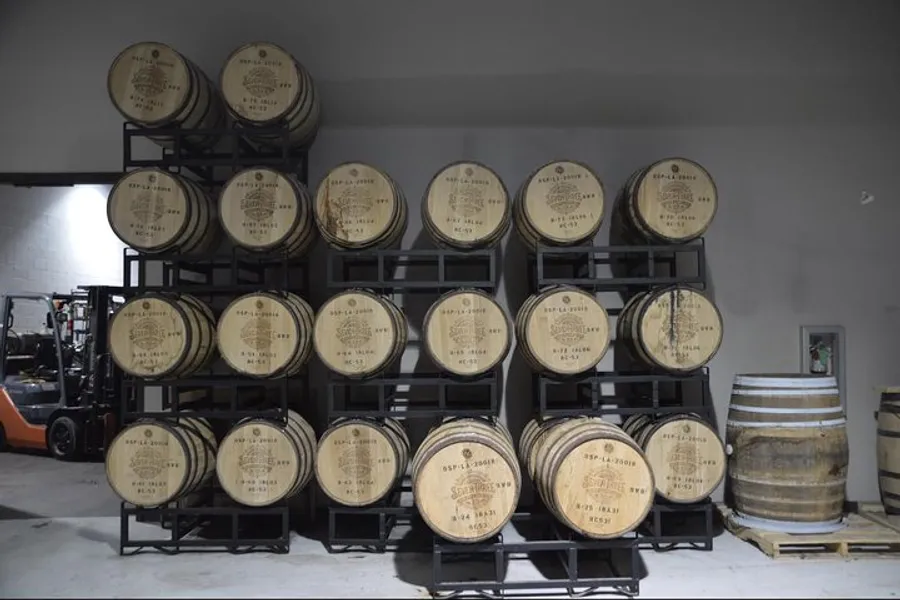 The image shows a storage area with several rows of wooden barrels, likely containing wine or spirits, stacked on metal racks.