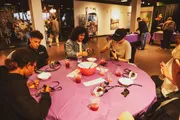 A group of individuals is seated around a table covered with a pink tablecloth, engaged in a craft activity with beads and string, with beverages nearby and other people in the background.