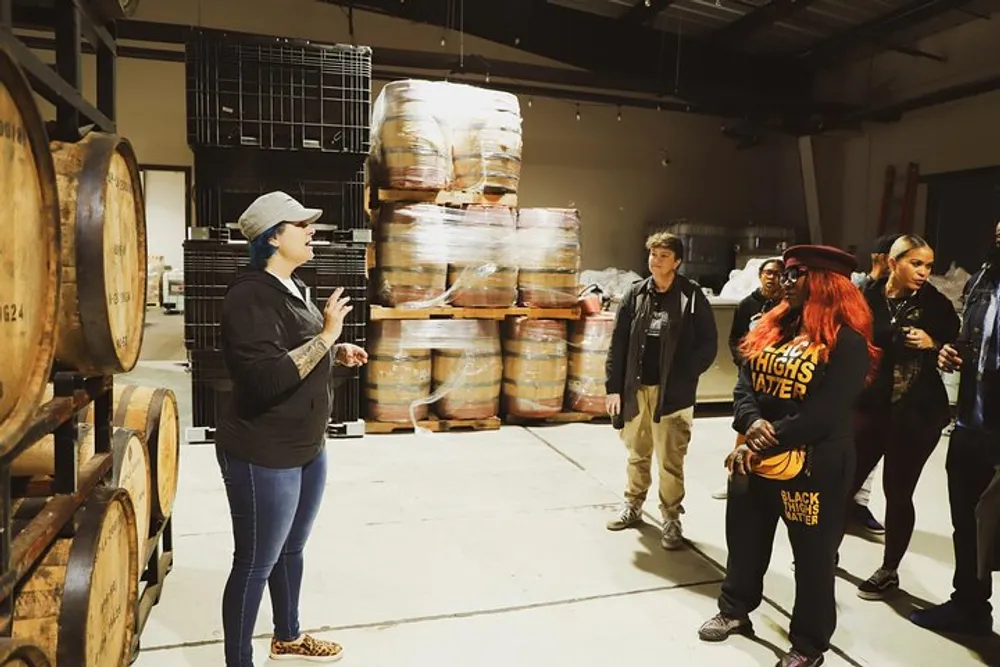 A guide is speaking to a group of people on a tour in a warehouse with stacked barrels and crates in the background