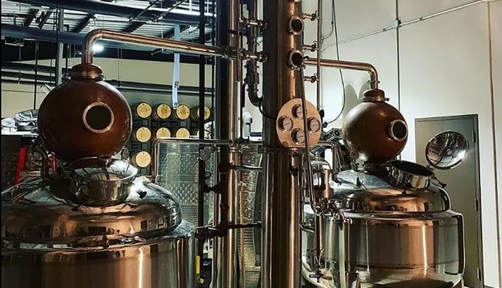 The image shows a copper distillation apparatus likely used for distilling spirits with various gauges and pipes in a facility with barrels visible in the background