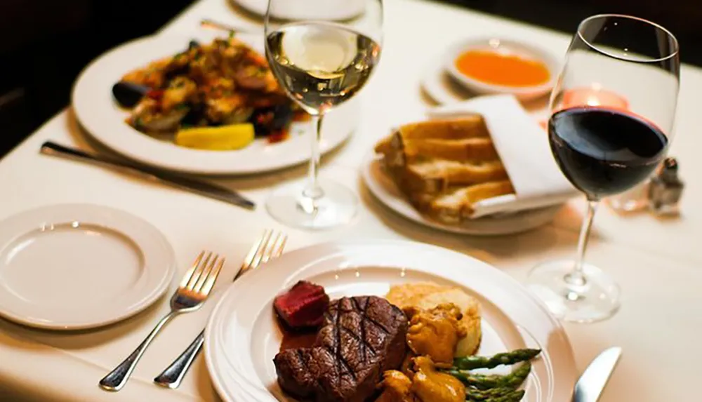 A dinner table is set with a plate of steak and vegetables in the foreground accompanied by a glass of red wine and a plate of mixed seafood with a glass of white wine in the background