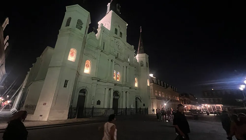 The image shows a historic cathedral lit up at night with people walking around the open square in front of it