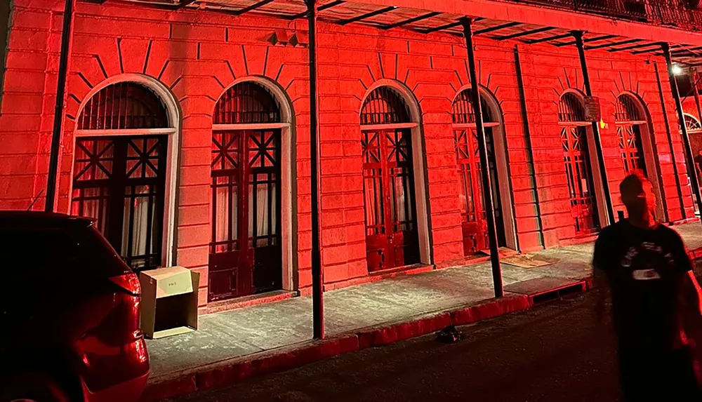 The image shows a historical building at night illuminated by red lights with arched doorways and columns a person in the foreground and part of a parked car on the left