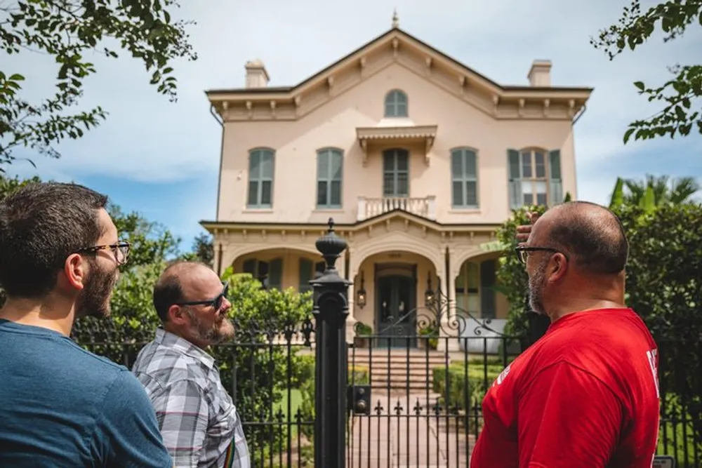 Three men are looking at an elegant two-story house from behind a wrought iron fence under a partly cloudy sky