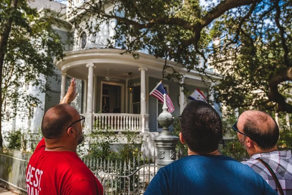 Three men are standing outside looking towards a classic white house with a wraparound porch and one of them is pointing up towards the second story