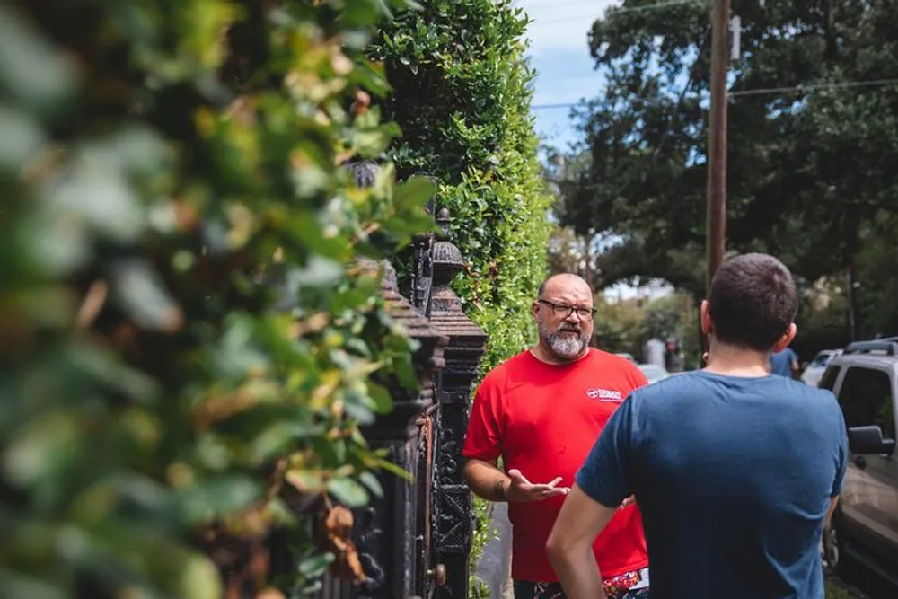 Two men appear to be engaged in a conversation by a fence lined with greenery with one facing the camera and the other with his back to the camera