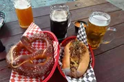The image shows a casual outdoor dining setting featuring a large pretzel, a loaded hot dog, and three different beers on a wooden table.